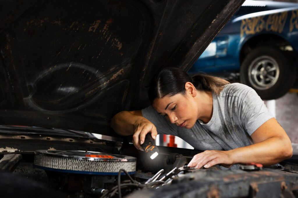 A mechanic, focused and using a flashlight, works on a car engine in an auto repair shop.