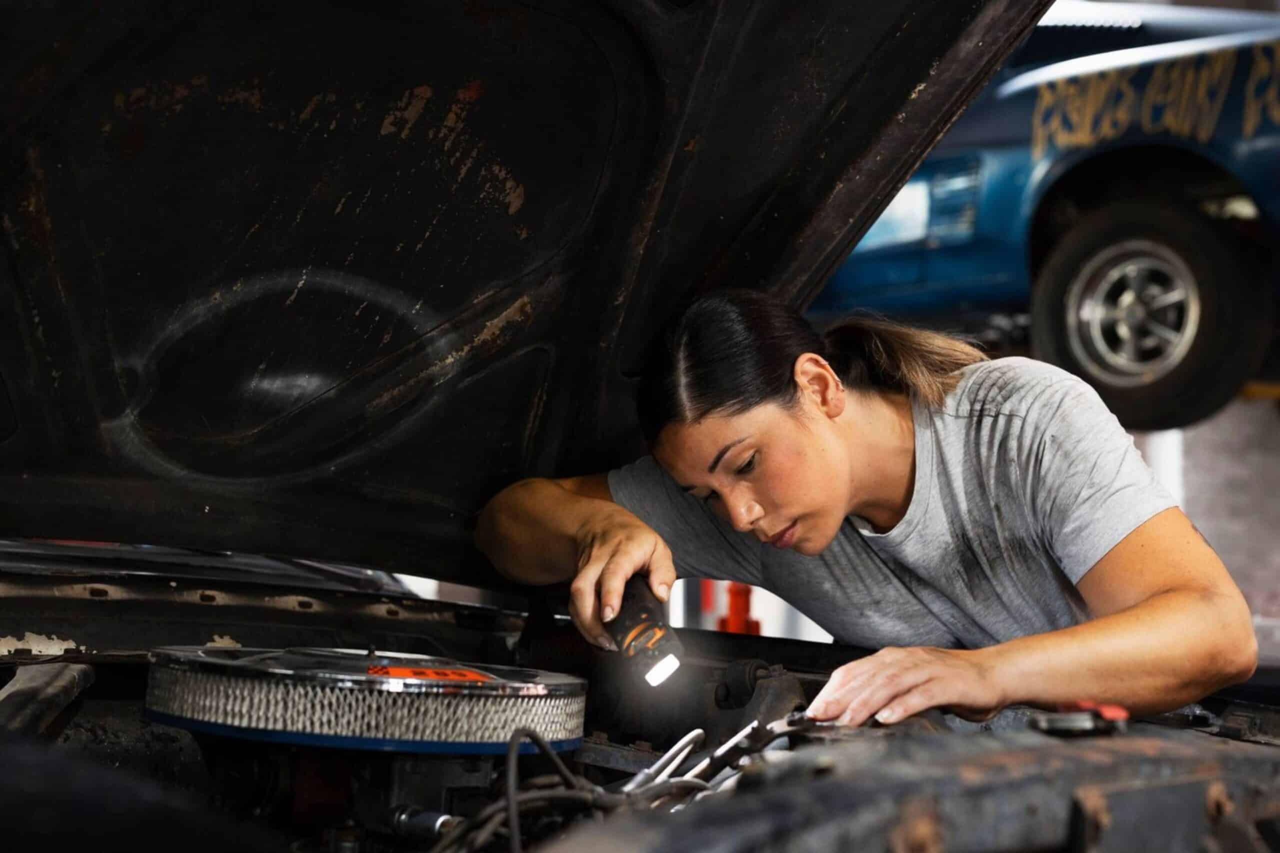 A mechanic, focused and using a flashlight, works on a car engine in an auto repair shop.