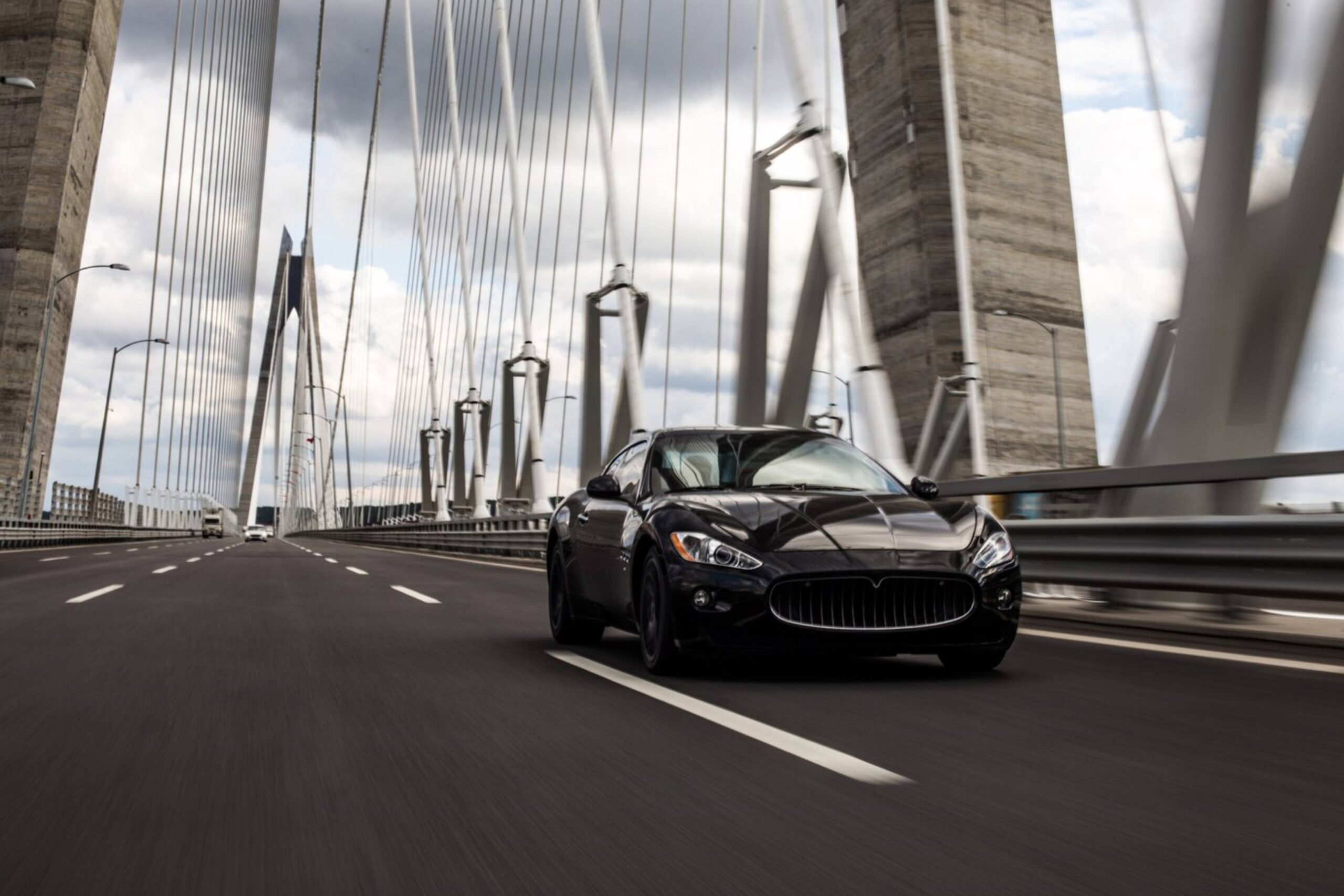 A black luxury car speeding on a multiple-lane suspension bridge with towering pillars and cables under a cloudy sky.