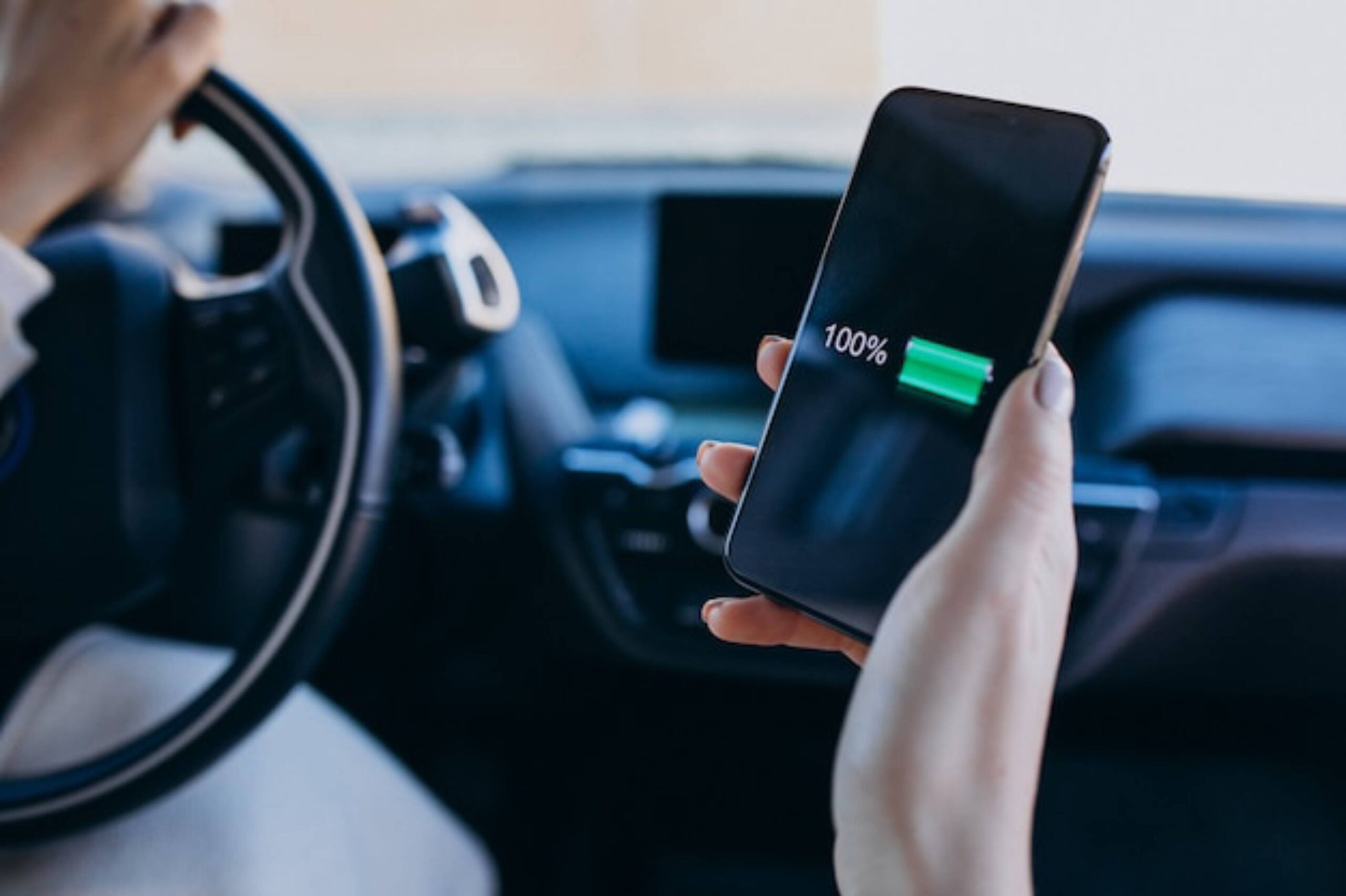  Driver in car holding smartphone with 100% battery charge displayed on screen.