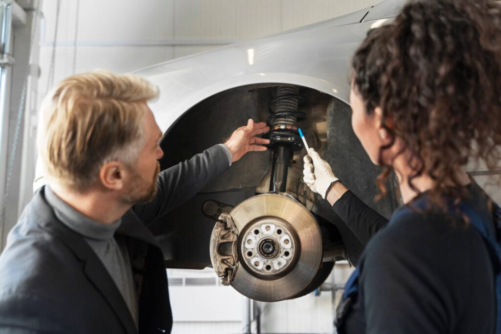 Two mechanics, a man and a woman, repair a car's suspension system in a workshop.