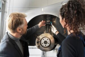 Two mechanics, a man and a woman, repair a car's suspension system in a workshop.