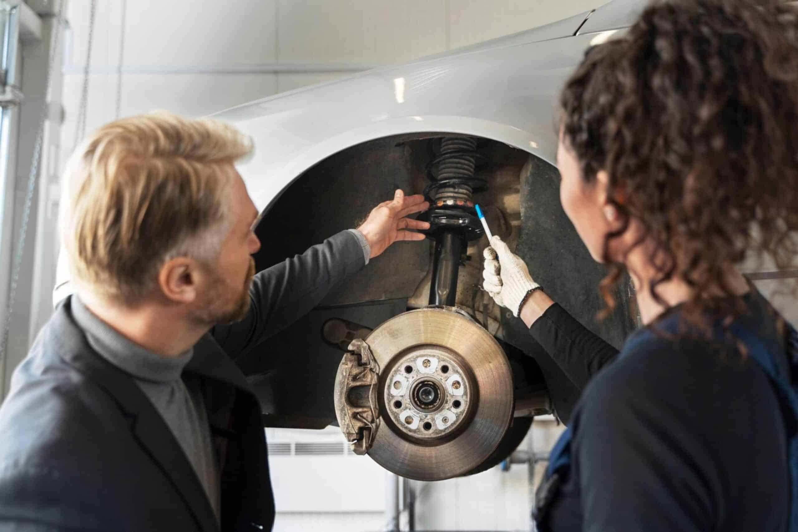 Two mechanics, a man and a woman, repair a car's suspension system in a workshop.
