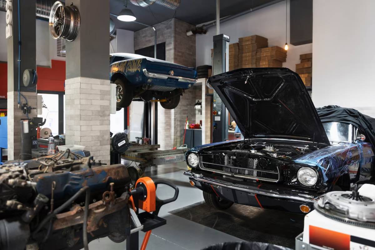  Classic cars being serviced in a busy, well-equipped garage with one car elevated on a lift and another with open hood.