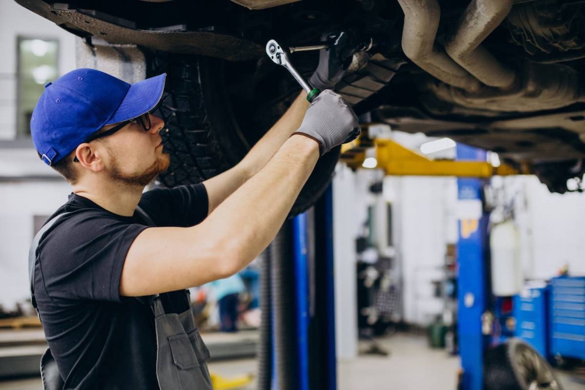  Mechanic in blue cap and gloves using a wrench on a car's underside in a workshop.
