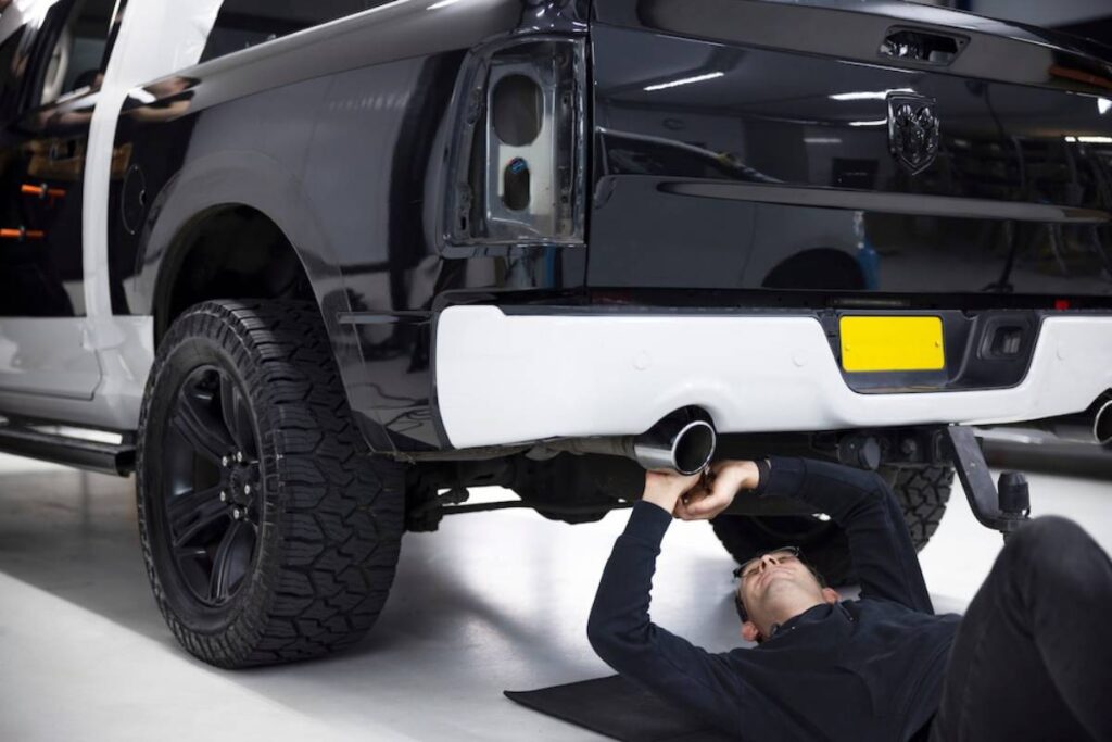 Mechanic lying on the floor while working underneath a raised, silver pick-up truck in a garage.