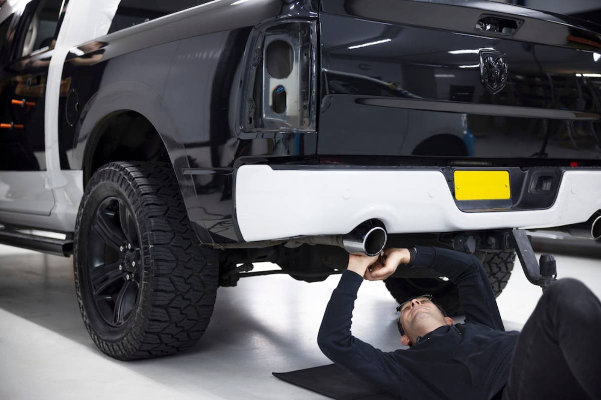 Mechanic lying on the floor while working underneath a raised, silver pick-up truck in a garage.