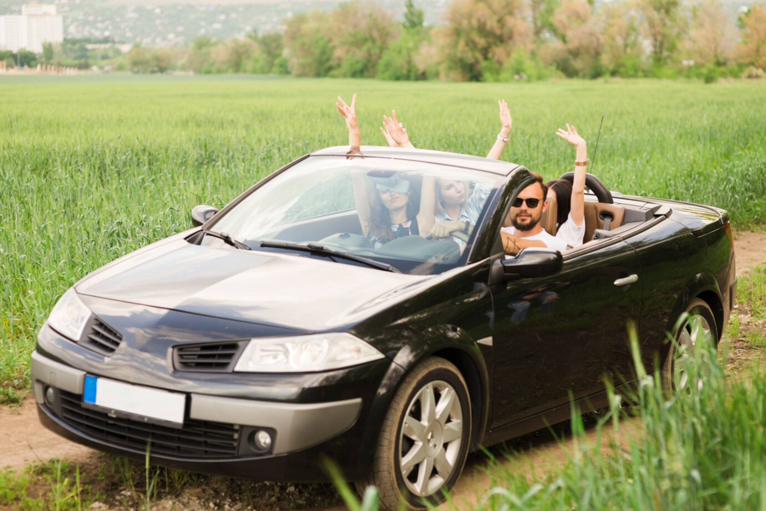 Three joyful friends in a convertible car waving in a green field. 