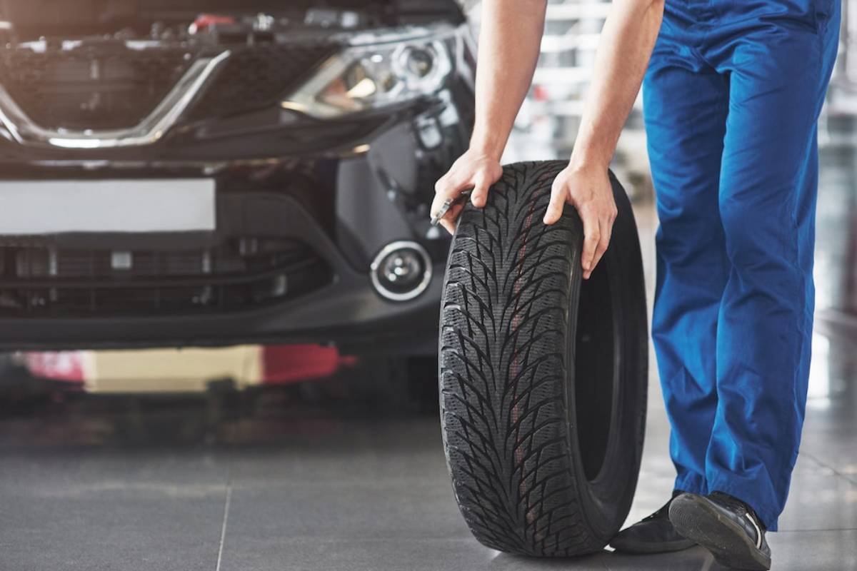  Mechanic holding a car tire in a workshop, with a vehicle's front visible in the background.